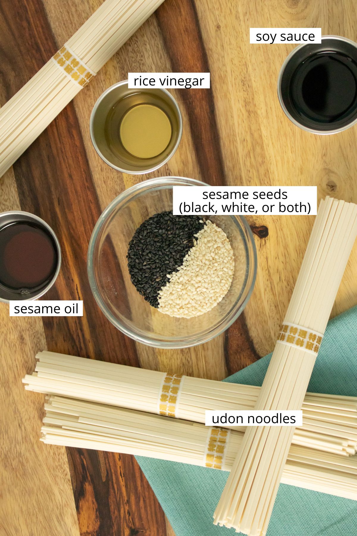 udon noodles on a wooden cutting board with ramekins of sesame seeds, sesame oil, rice vinegar, and soy sauce, all labeled with text