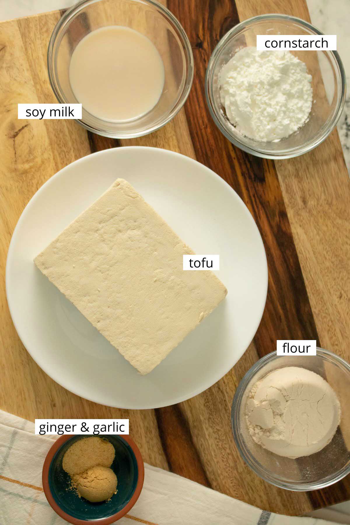 tofu, cornstarch, flour, soy milk, and spices arranged on a wooden cutting board with text labels