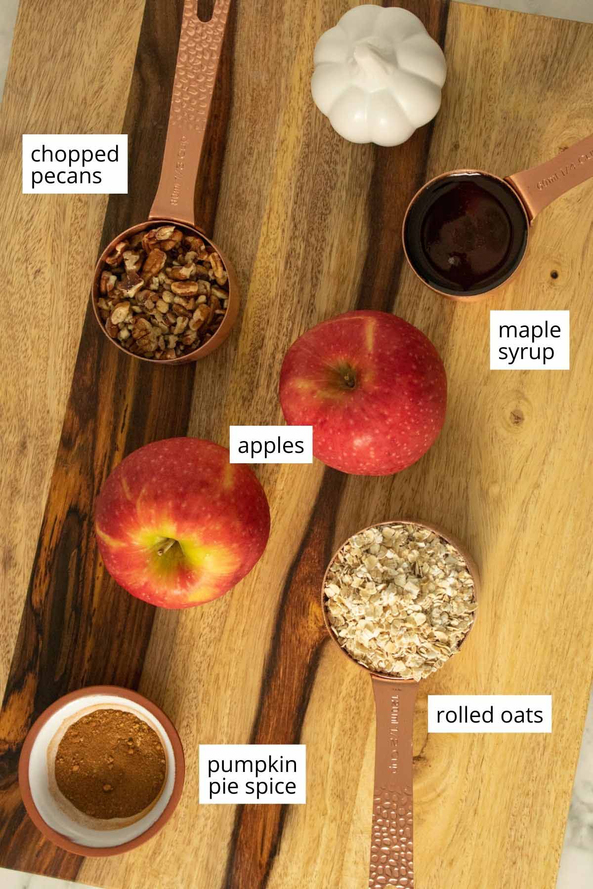 apples, maple syrup, oats, pumpkin pie spice, and pecans on a wooden cutting board. Text labels on each ingredient