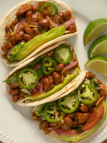 3 pinto bean tacos with veggie toppings on a white plate with limes