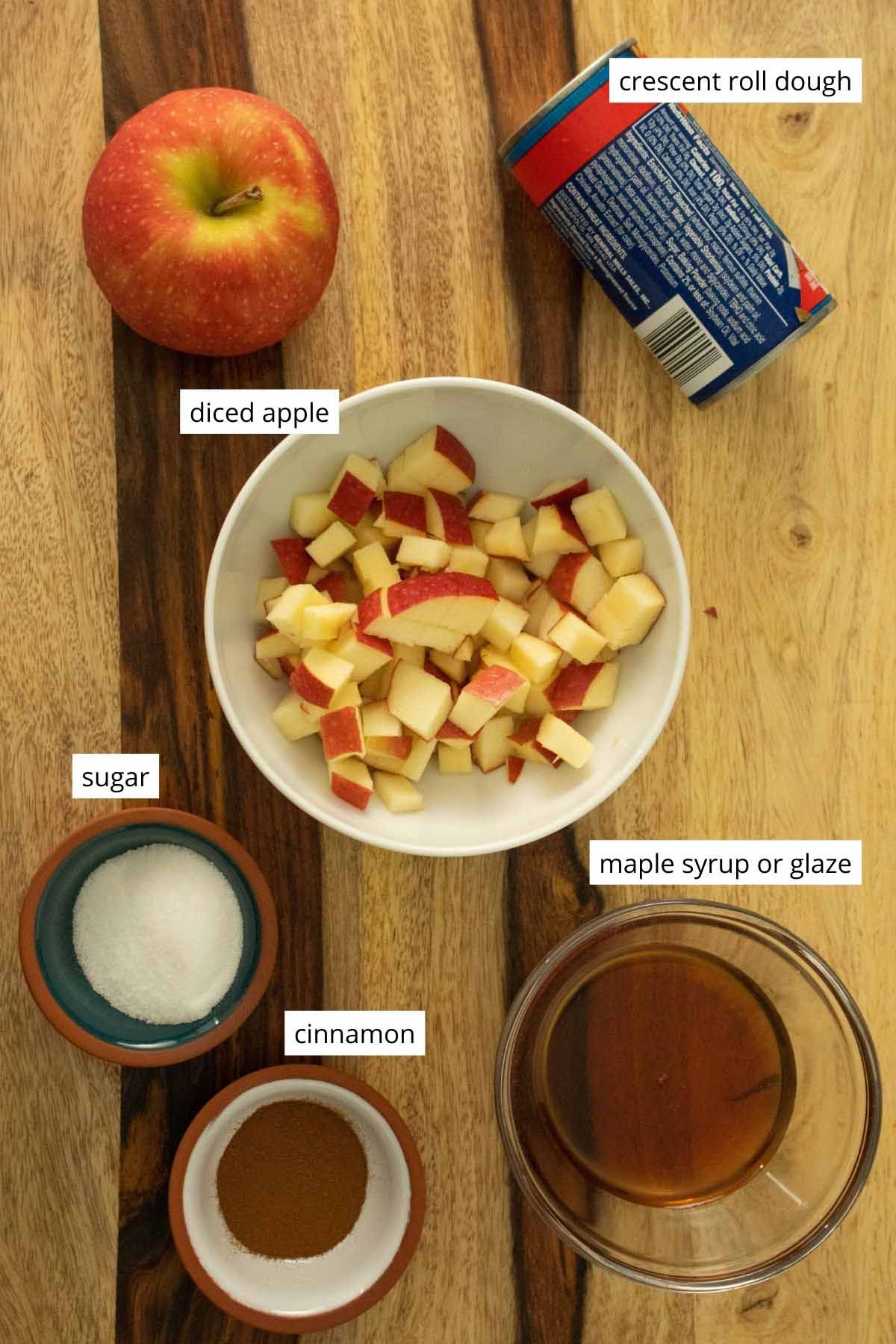 photo of diced apples, crescent roll dough package, cinnamon, sugar, and maple syrup all on a wooden cutting board