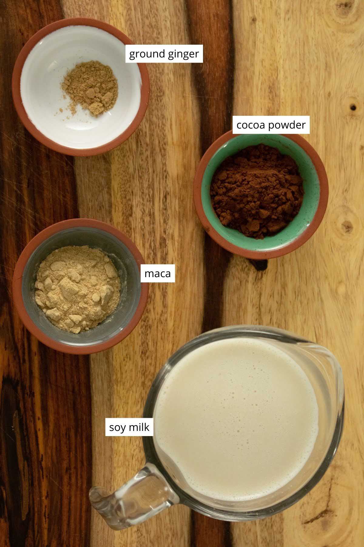 powdered ginger, maca, and cocoa in cups on a cutting board next to a measuring up of soy milk