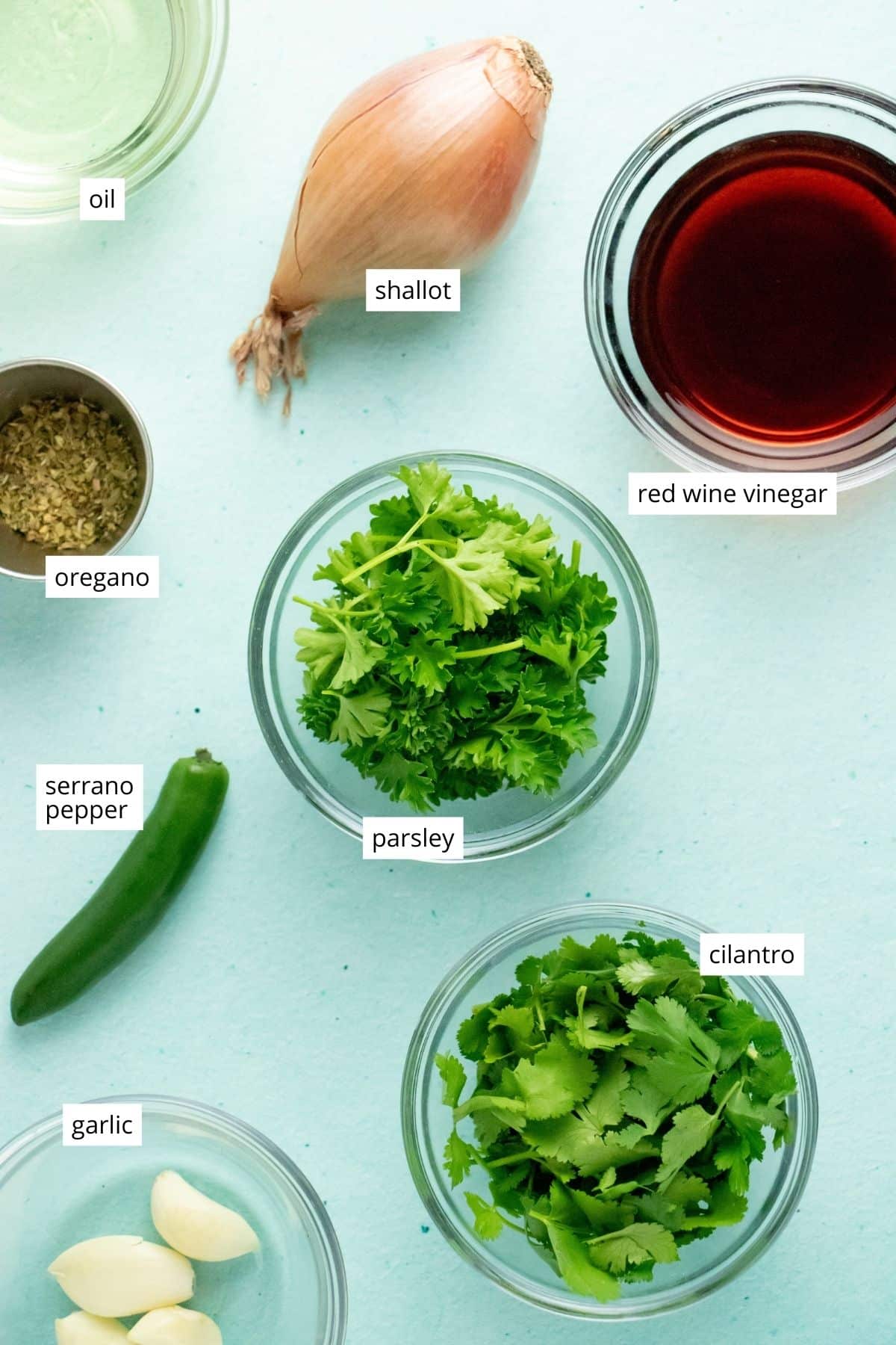 cilantro, parsley, and other chimichurri ingredients in bowls on a blue table