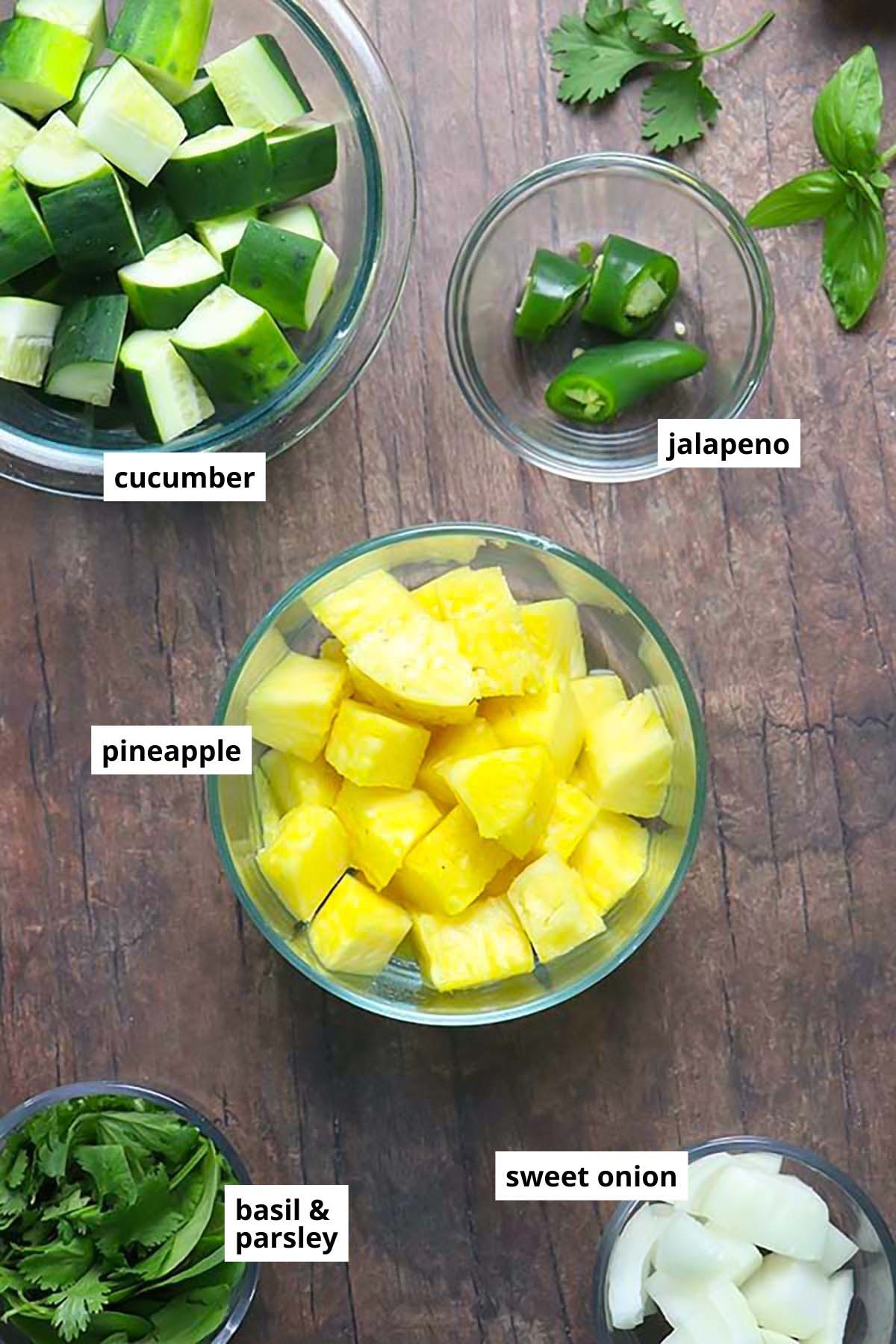 pineapple, cucumber, herbs, onion, and jalapeno in bowls on a wooden table with text labels