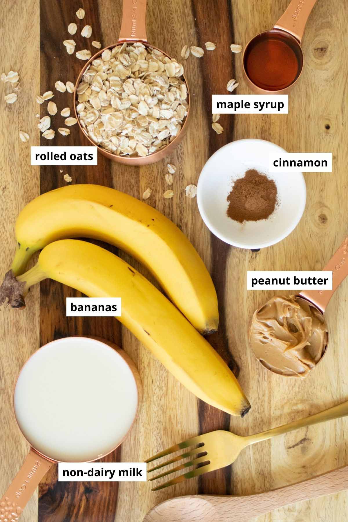 bananas, peanut butter, rolled oats, soy milk, cinnamon, and maple syrup in measuring cups on a wooden table