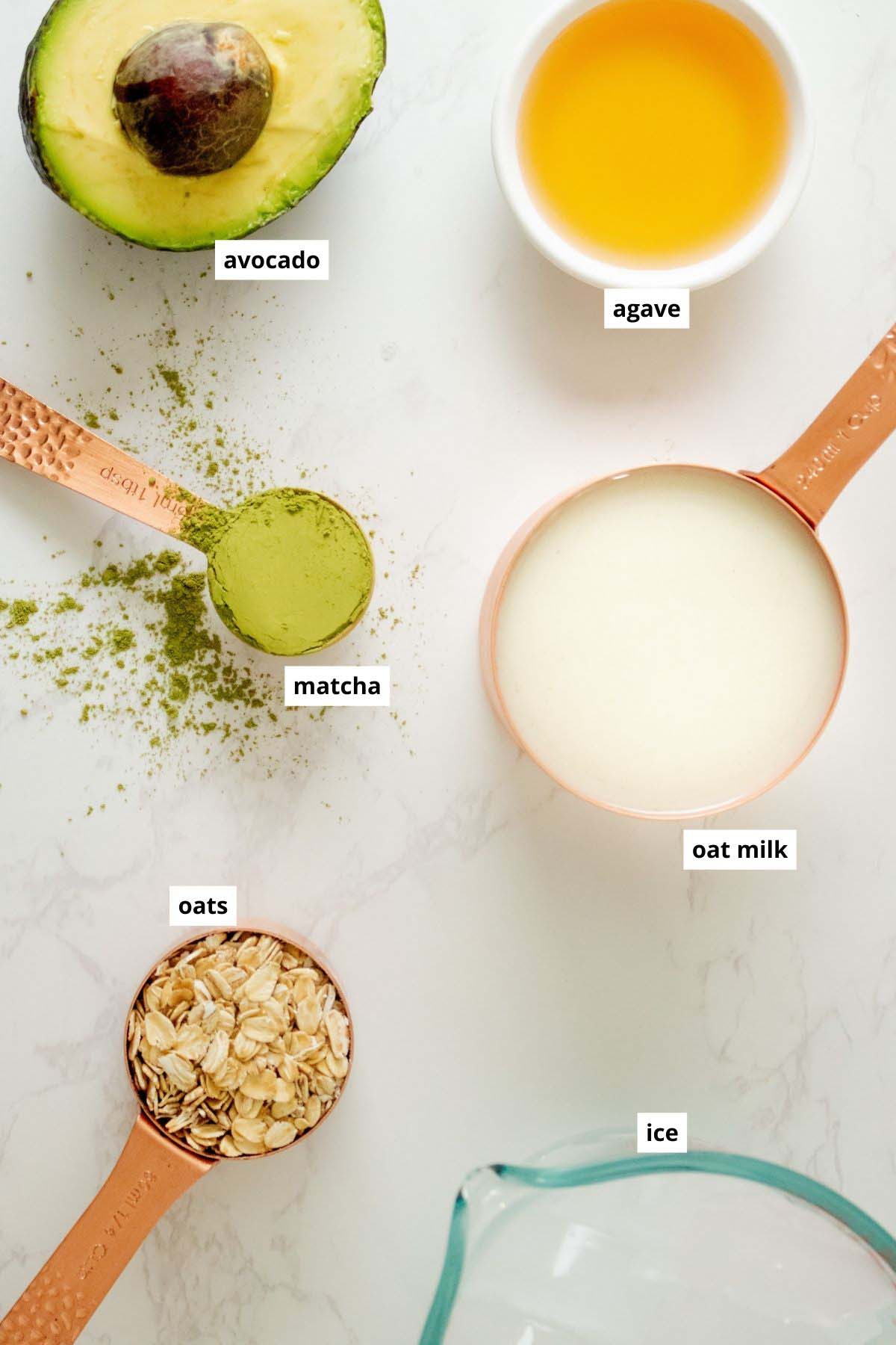 matcha powder, avocado, agave, oat milk, ice, and oats in cups on a white table with text labels on each one