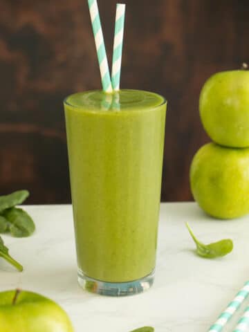 green apple smoothie in a glass with apples and spinach on the table around it