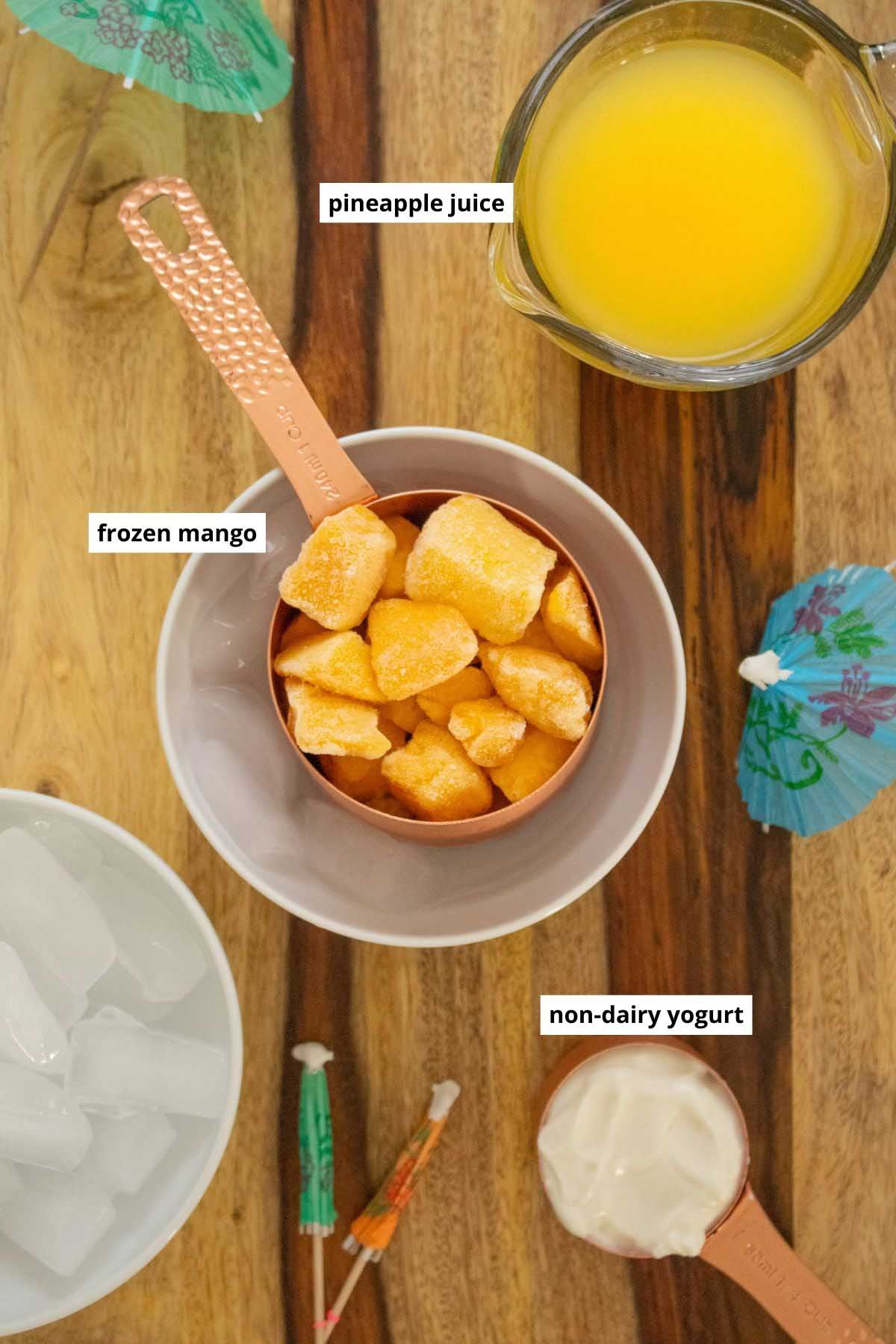 frozen mango, pineapple juice, non-dairy yogurt, and ice in bowls on a wooden countertop