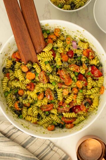bowl of pesto pasta salad with veggies in a white serving bowl and wooden serving spoons