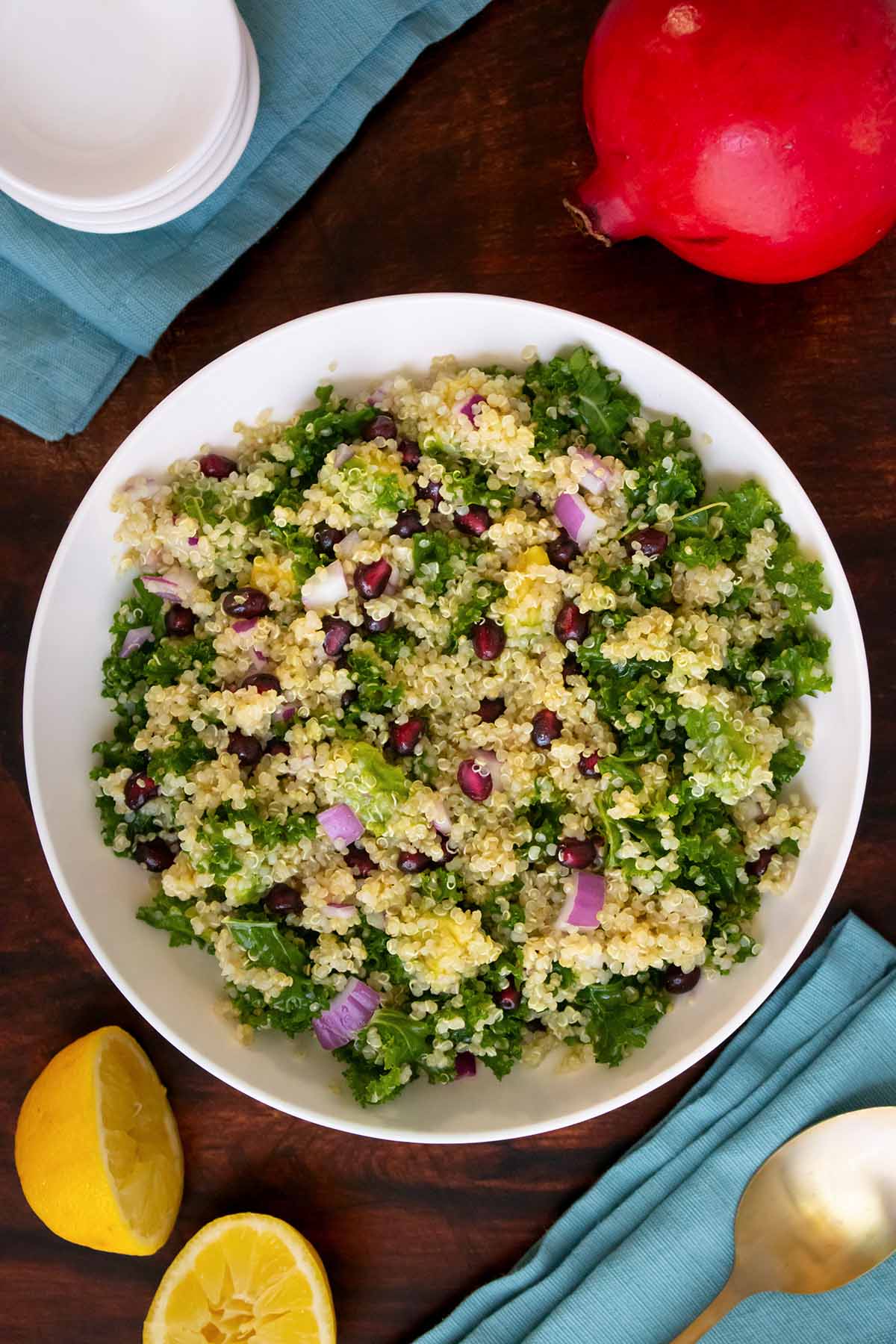 bowl of quinoa salad with kale, pomegranate seeds, avocado, and red onion on a table with fruits and napkins