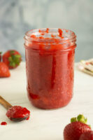jar of low sugar strawberry freezer jam on a white table surrounded by strawberries and a spoon with the jam in it