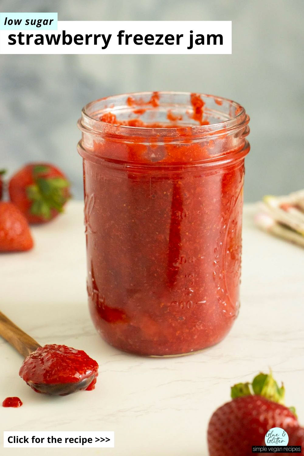 jar of low sugar strawberry freezer jam on a white table surrounded by strawberries and a spoon with the jam in it, text overlay