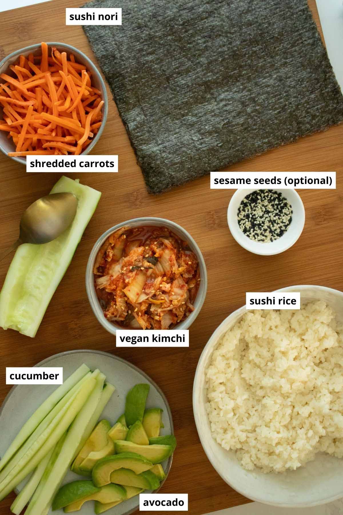 kimchi, sushi rice, nori sheets, sesame seeds, and veggies arranged on a wooden cutting board
