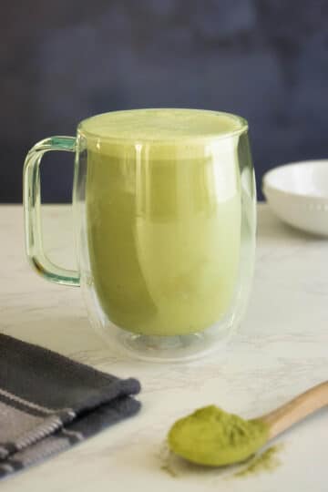 vegan matcha latte in a glass mug with a spoonful of matcha powder next to it