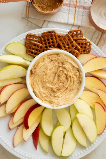 whipped peanut butter yogurt dip topped with cinnamon on a plate with pretzels and apples