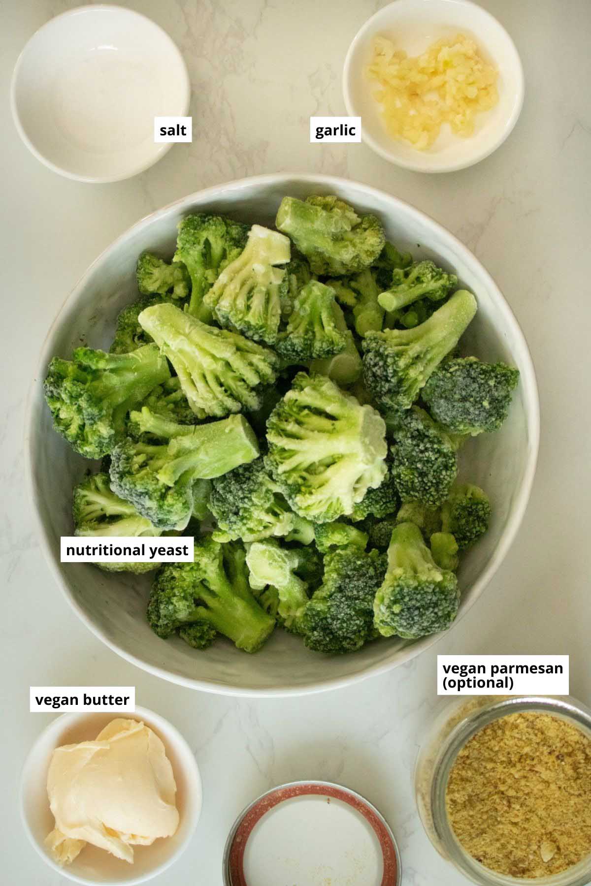 frozen broccoli, garlic, vegan butter, vegan parmesan, and salt in bowls on a white table