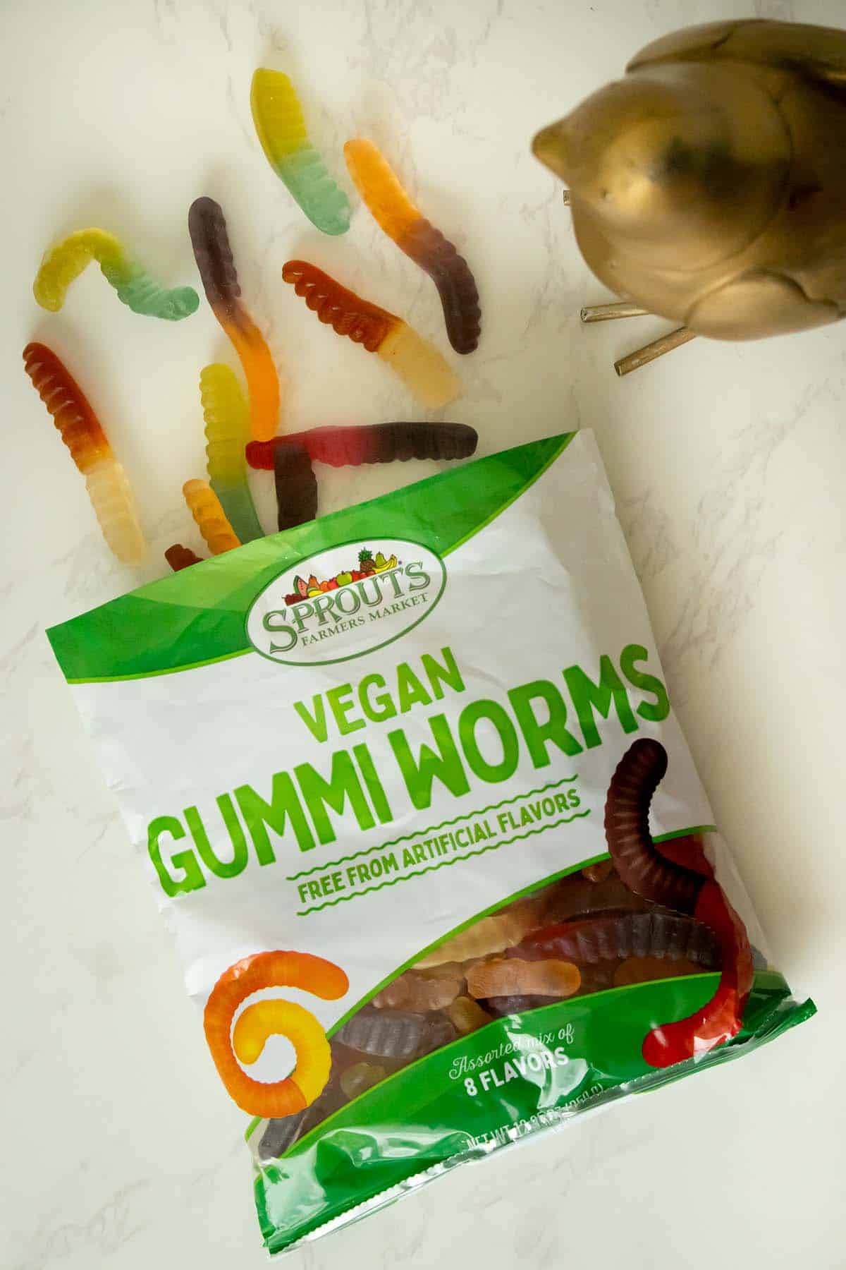 a bag of Sprouts Vegan Gummi Worms on a white table next to a brass bird