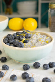 lemon blueberry chia pudding in a bowl on a white table with lemons, berries, and chia seeds around it
