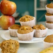 banana apple muffins on a white plate surrounded by apples, banana, and a muffin broken in half, so you can see the texture inside