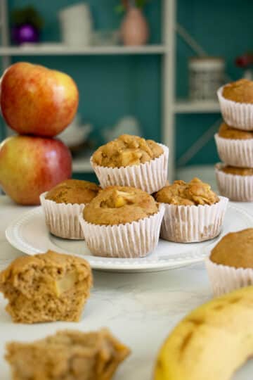 banana apple muffins on a white plate surrounded by apples, banana, and a muffin broken in half, so you can see the texture inside