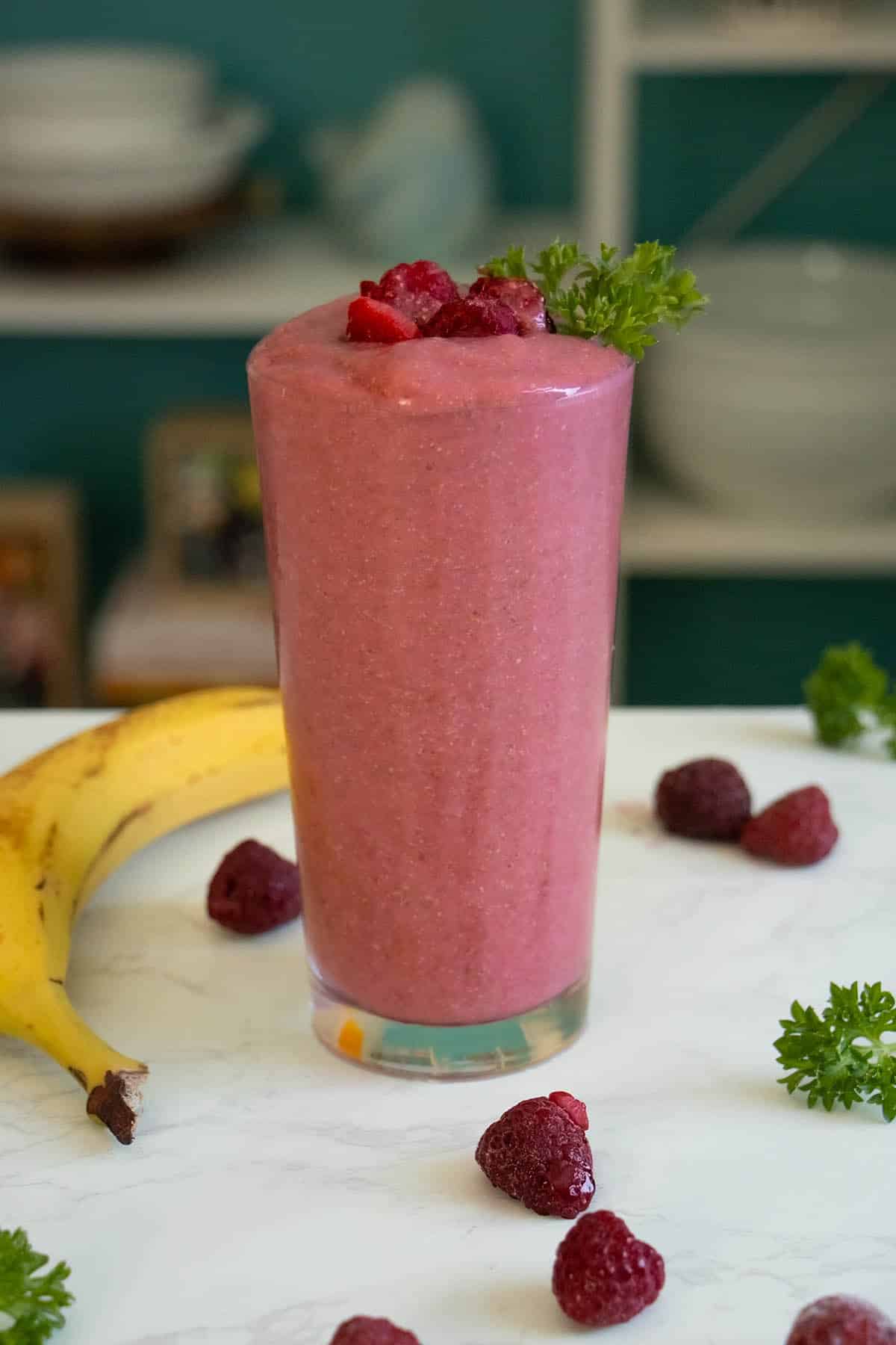 banana and raspberry smoothie in a glass on a white table surrounded by fruit and parsley