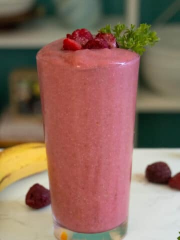 banana and raspberry smoothie in a glass on a white table surrounded by fruit and parsley