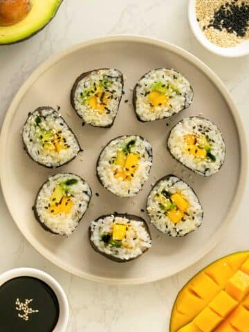 mango roll sushi on a plate with sesame seeds surrounded by soy sauce, mango, avocado, and a cupful of sesame seeds