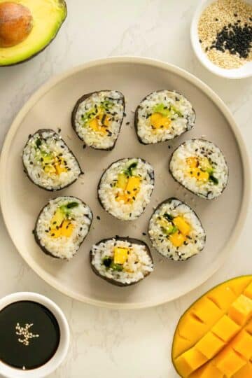 mango roll sushi on a plate with sesame seeds surrounded by soy sauce, mango, avocado, and a cupful of sesame seeds