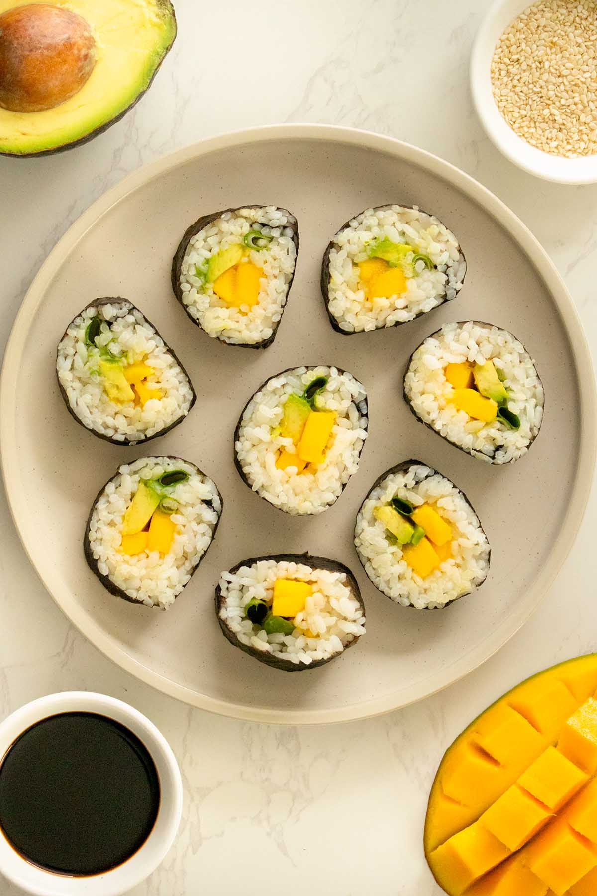 mango roll sushi on a plate before adding sesame seeds surrounded by soy sauce, mango, avocado, and a cupful of sesame seeds, text overlay