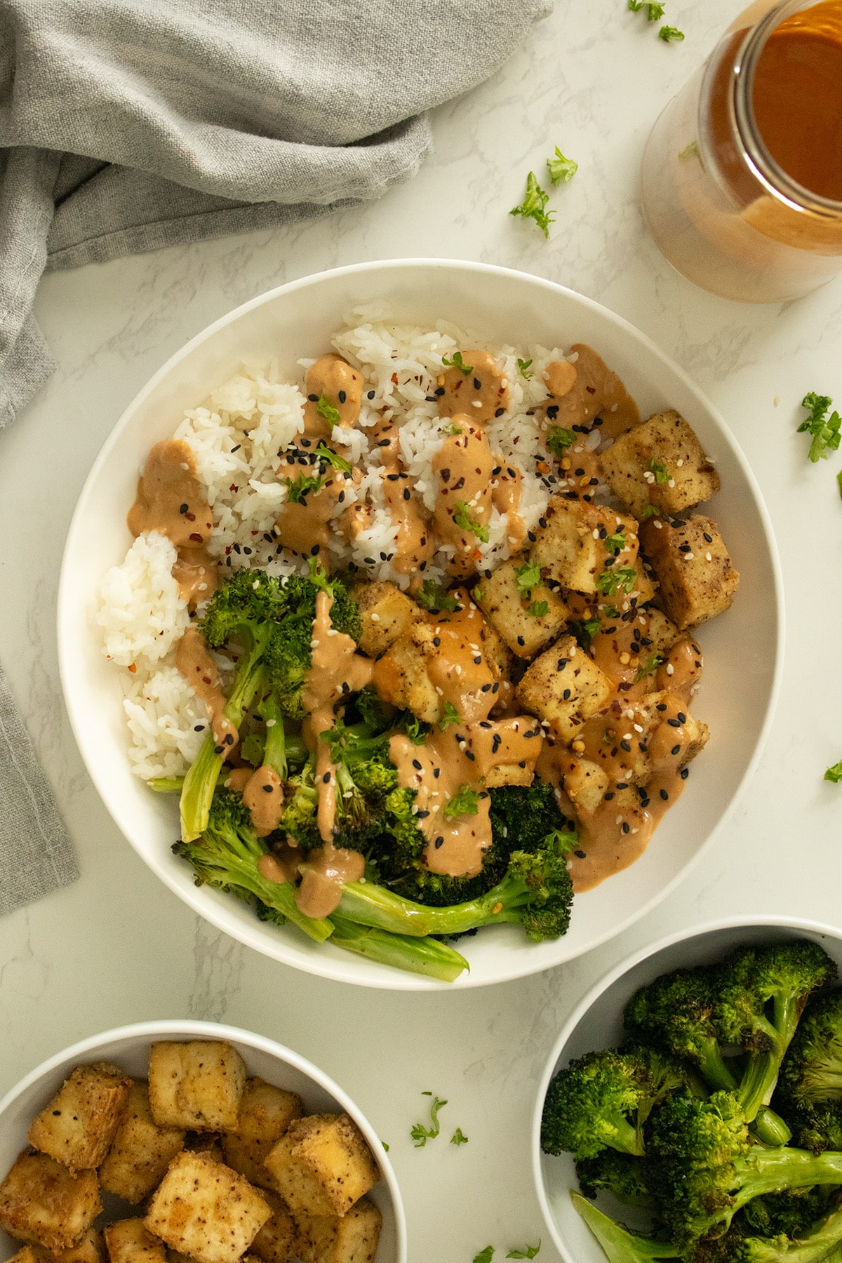 baked tofu in peanut sauce in a white bowl with rice, bowls of tofu and broccoli and a jar of more sauce on the side