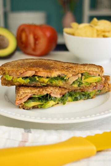 grilled avocado sandwich on a white plate with avocado, tomato, and chips in the background