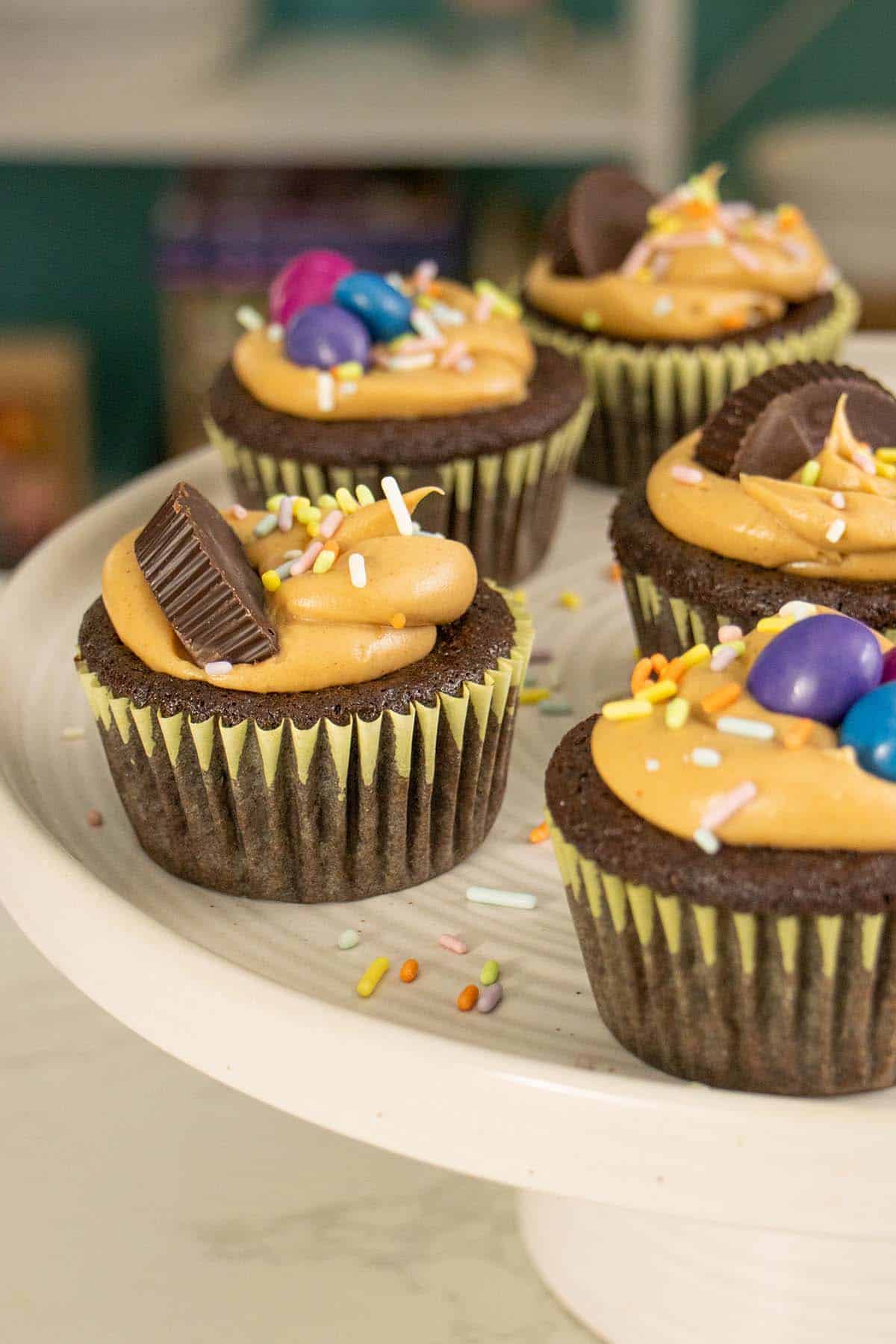 chocolate peanut butter cupcakes on a cake stand decorated with frosting, sprinkles, and candy