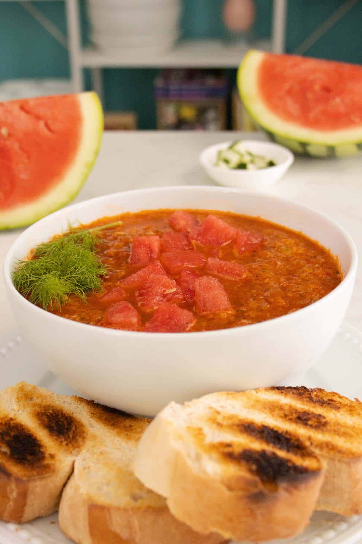 bowl of watermelon gazpacho with toast in the foreground and big watermelon slices in the background