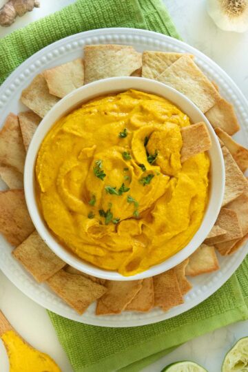 carrot hummus in a serving bowl with pita chips and ingredients around it and a pita chip dipped into the hummus