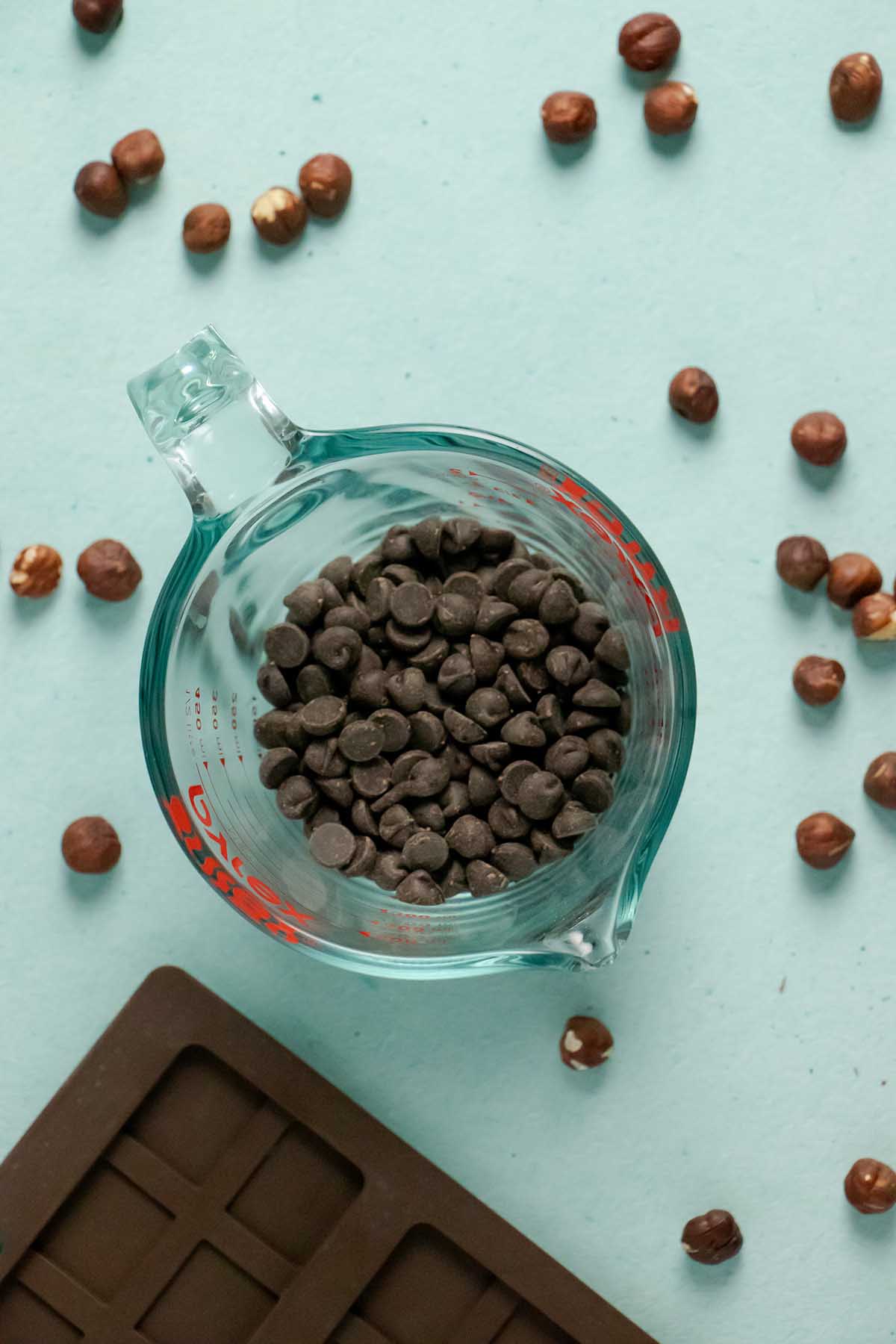 vegan chocolate chips in a measuring bowl on a table with hazelnuts and the chocolate mold