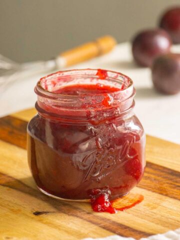 jar of plum freezer jam on a wooden cutting board with plums in the background
