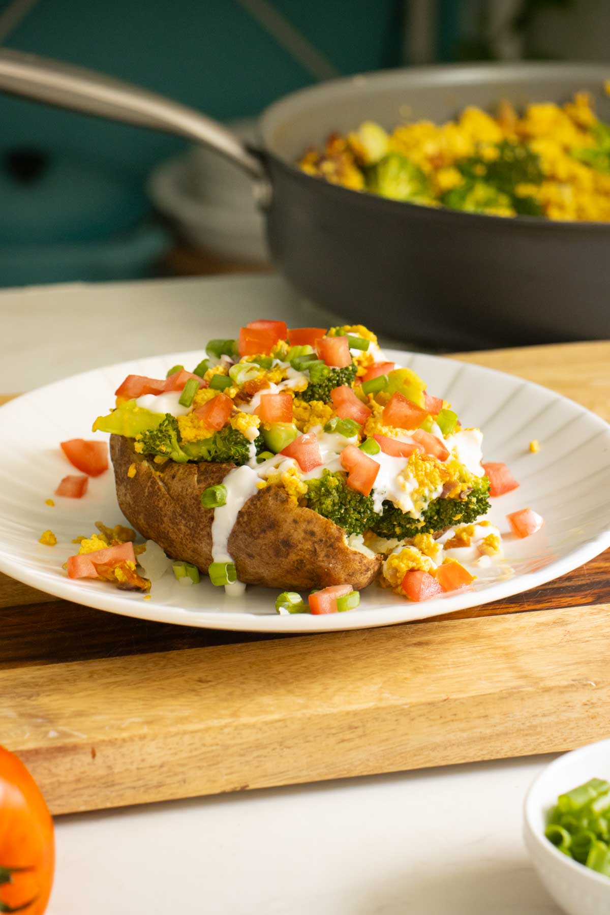 loaded baked breakfast potato on a plate with all the toppings, pan of tofu scrambled the background