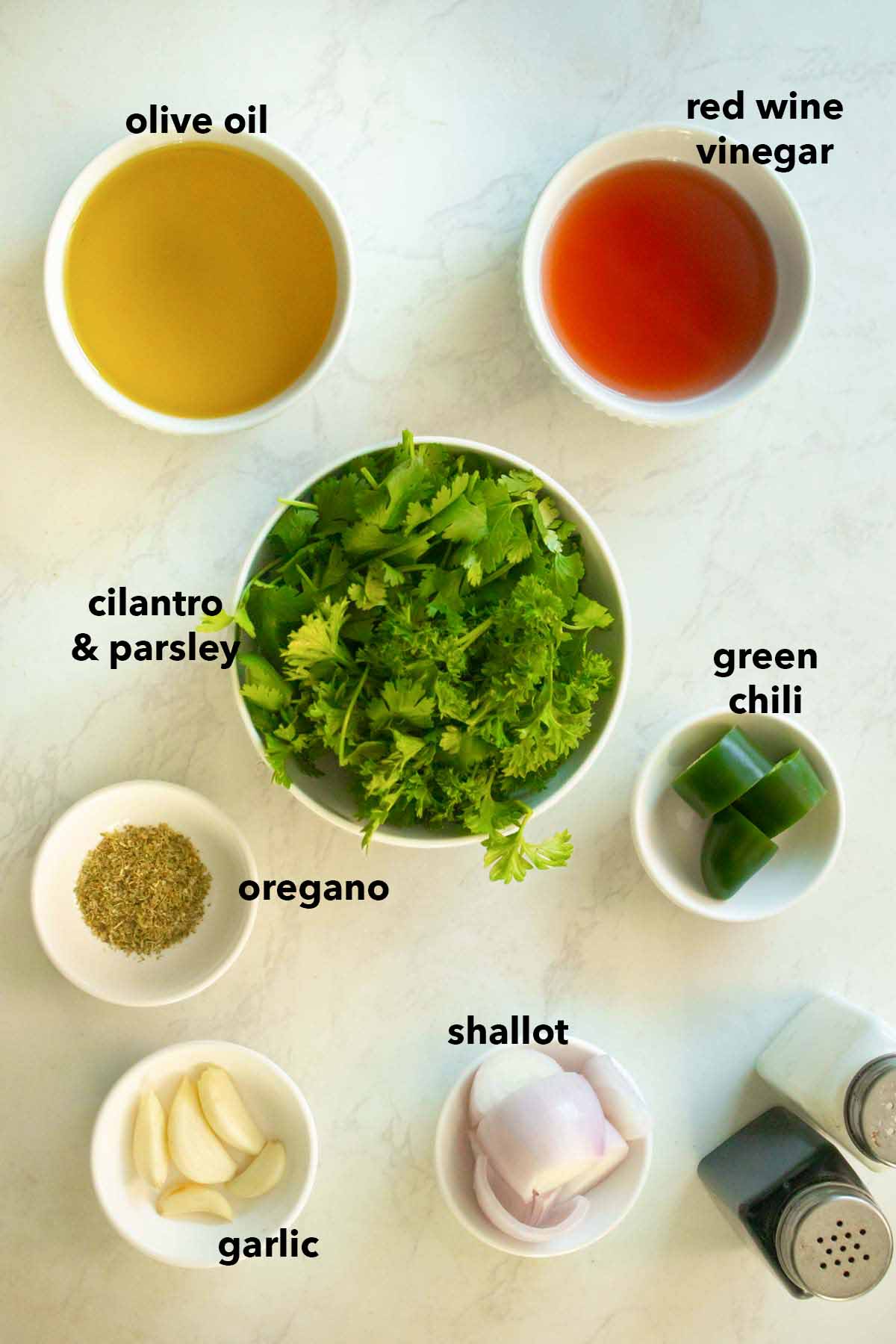 chimichurri ingredients in white bowls on the kitchen counter with text labels on each ingredient